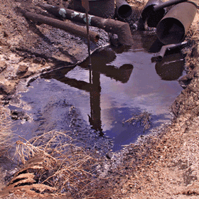 A photo of rusty pipes draining dirty water into a small, muddy puddle