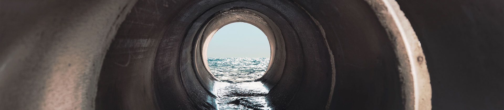 Inside view of a long effluent sewer pipe with a body of water visible at the mouth of the pipe.