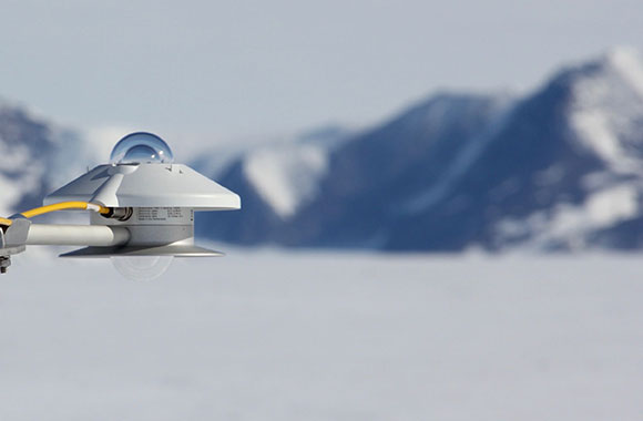 A Kipp & Zonen pyranometer in the foreground is mounted to a bracket. A blurred Background includes a snow-covered field and mountain range.